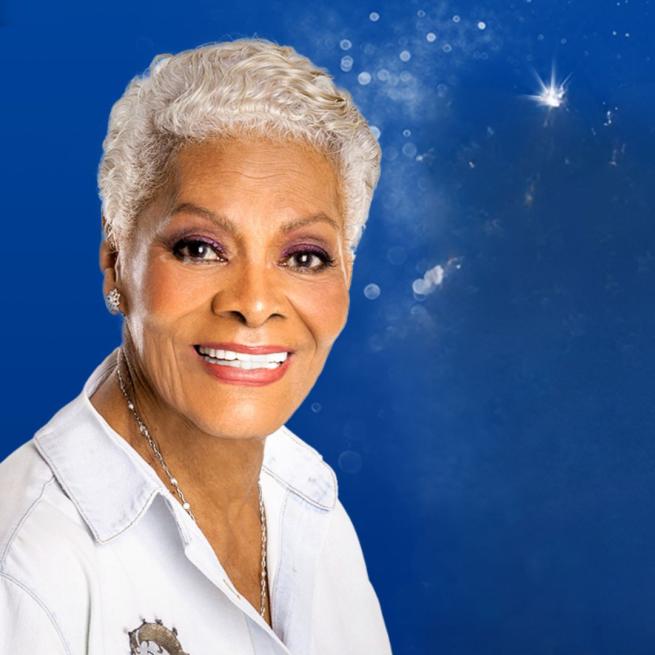 Dionne Warwick smiling at the camera wearing a white shirt with an image of Betty Boop on a motorbike stitched into it.