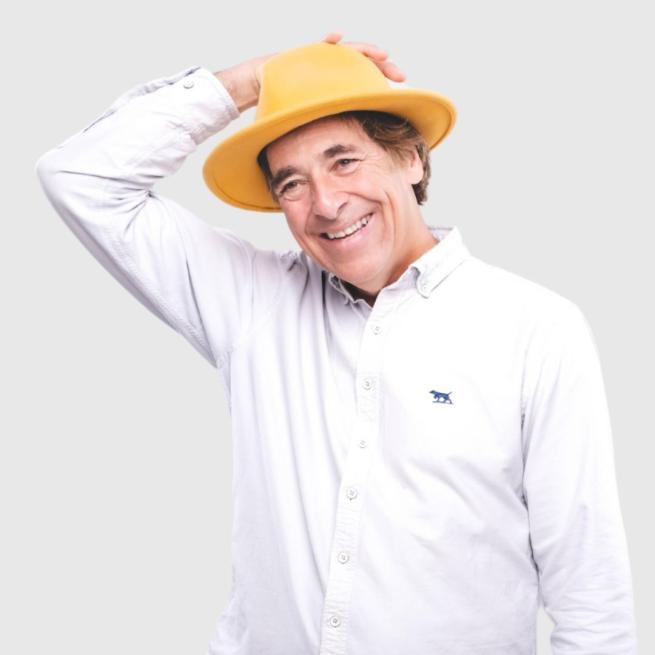 Mark Steel in a white shirt wearing an orange-yellow hat. His right hand is holding the hat on his head and he is smiling.