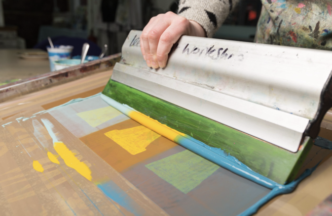 A squeegee pulling green, yellow and blue inks across a screen with a design featuring three plant pots.