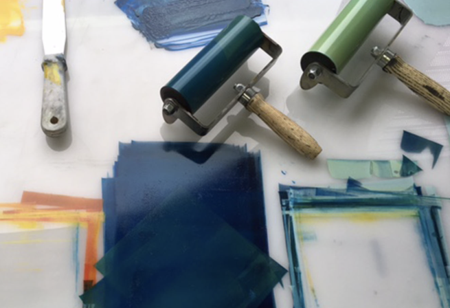 A pallette knife and two printmaking rollers with green and blue inks being rolled on a white surface.