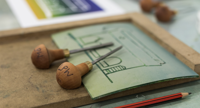 A piece of lino with an image of a building cut into it, together with some lino cutting tools.