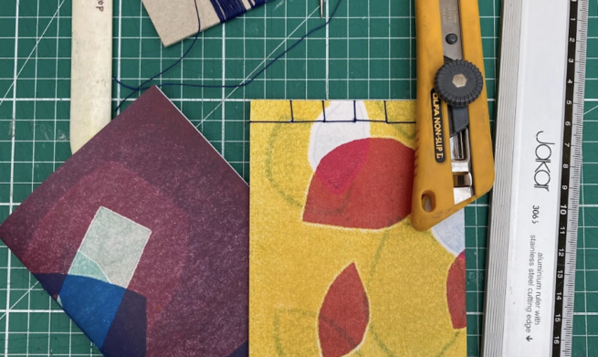 A selection of bookbinding tools on a cutting mat, including a craft knife, bone folder, cotton and a steel ruler. They are with some hand-bound books with abstract monoprinted covers in multiple colours.