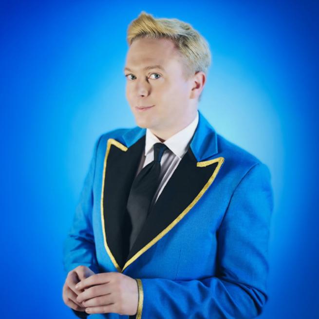 Stephen Bailey in an electric blue suit with a black tie and white shirt smirking against a blue background.