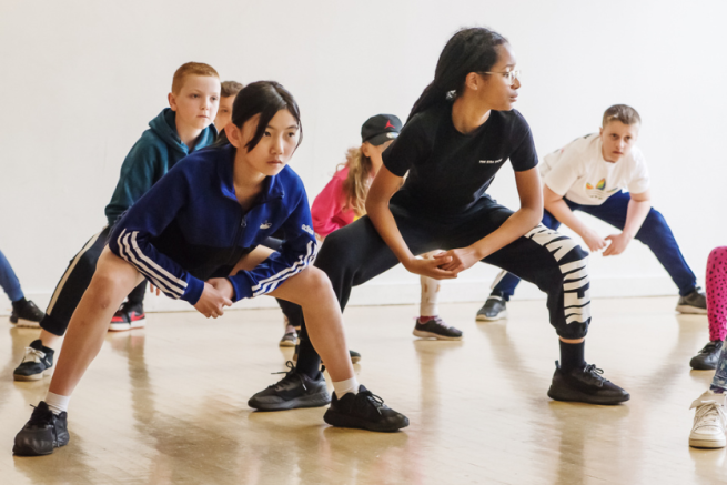 Children in a squat position looking to the left, the two children at the front are wearing dark gym clothes and the room is full 