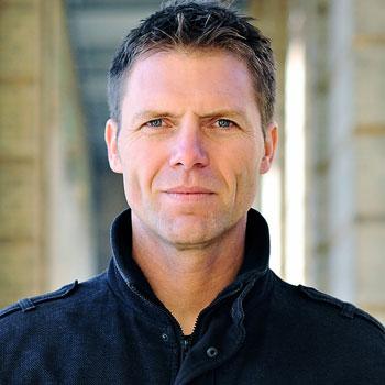 Middle aged white man with short brown hair and blue eyes looking into the camera with a stern face. He's wearing a dark coat with a big collar.