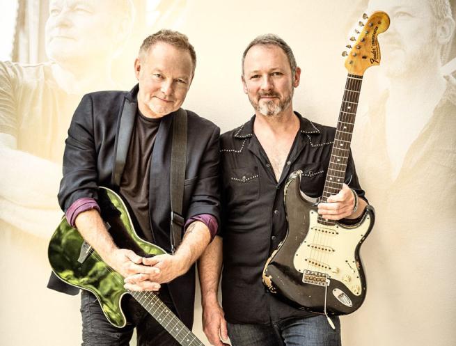 Two men holding electric guitars, standing side by side against a light background with subtle reflections of their images layered in the design. One wears a dark blazer and the other a black shirt with white accents.