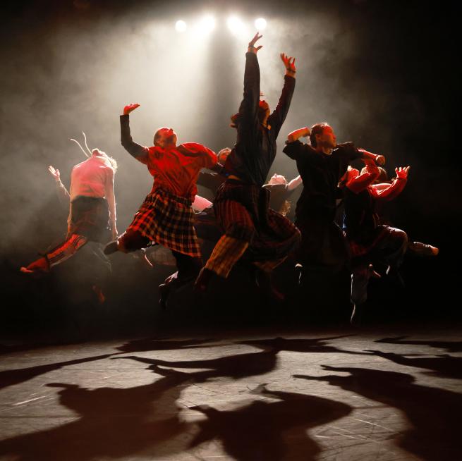 A group of dancers jump in the air. They are lit in red and white lighting. There are shadows reflected onto the stage.