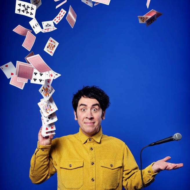 Pete Firman in a mustard-coloured shirt firing a pack of playing cards into the air with his right hand, his left levitating a cord microphone above it.