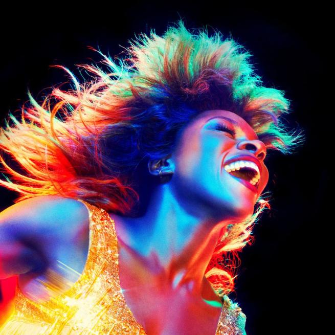 Image of Tina Turner with a rainbow tinge to the image. Tina is smiling wide in a gold fringe dress