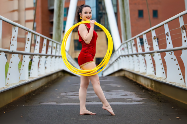 A woman in a red leotard stands with a yellow hula hoop on a bridge 