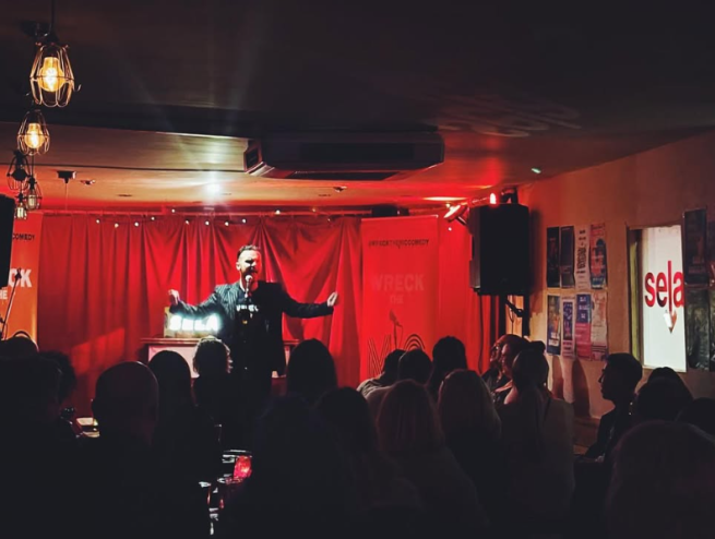 A white man with a black pin stripe suit and brown hair stands with his arms open on a stage in a red lit room, infront of a crowd of people.