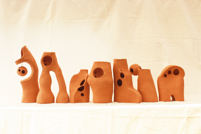 A row of abstract terracotta sculptures with organic shapes, rounded edges, and holes, displayed against a neutral fabric backdrop.