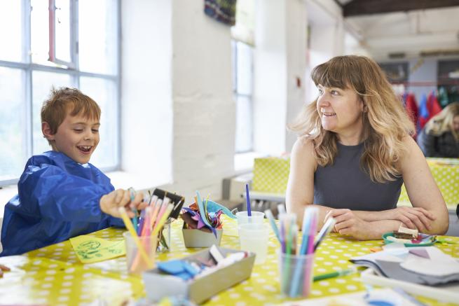 A child laughs, using paint and rollers, sitting next to a smiling adult.