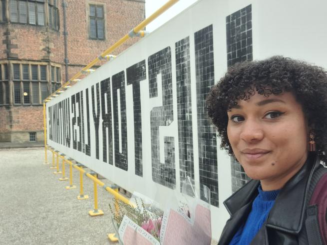 A women with dark curly hair stands in front of a big poster 