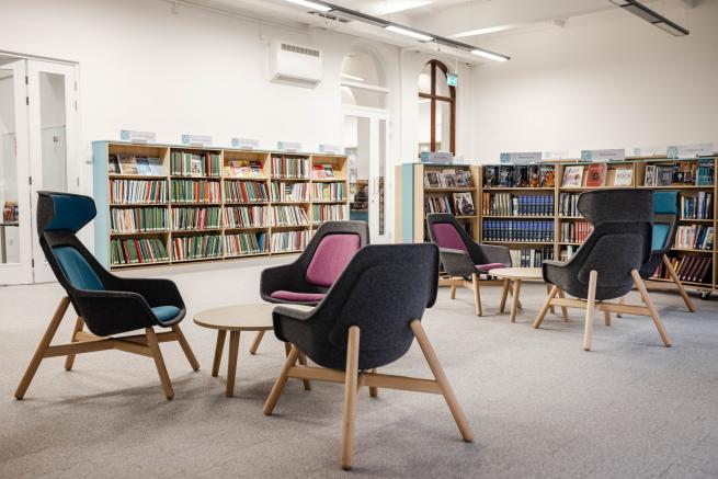 Picture of the music library. White walls with ash effect book shelves lining the room full of music books. Centre picture 6 chairs with Ash wood effect legs, the seat comprising of grey fabric with magenta and teal cushioning. The room is carpeted with a grey carpet.