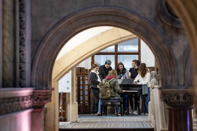 Group of six people stood round a piano in Leeds central library. Seen through the libraries majestic archways and stone work.