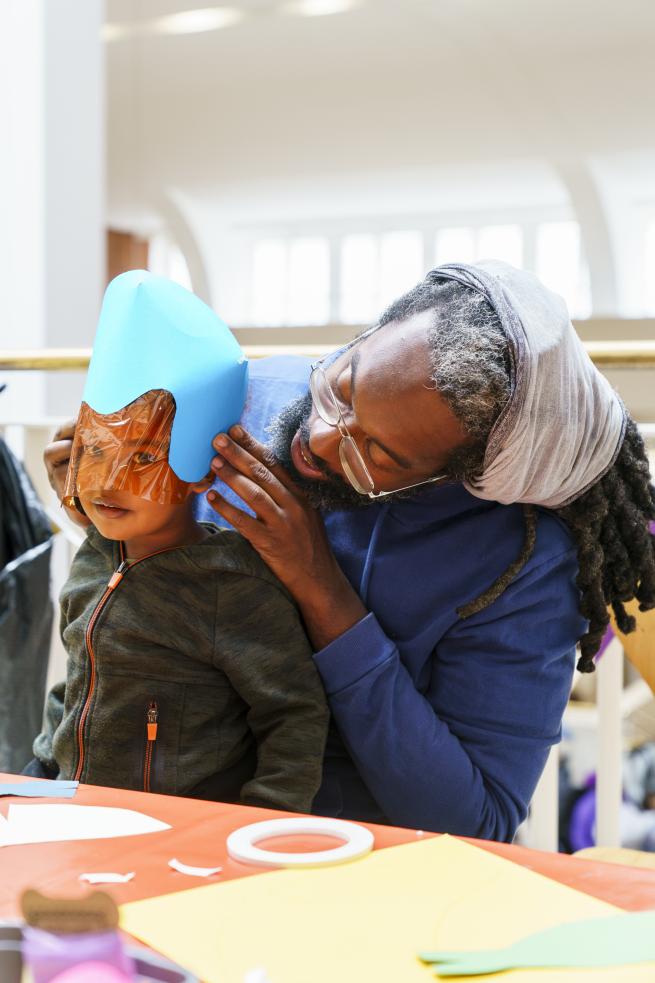 A father helps his child make a mask from card and transparent plastic.