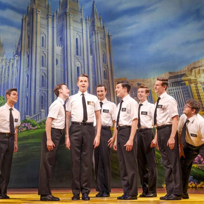 Cast of the Book of Mormon in their white shirts, black ties and black trousers singing. The Church of Jesus Christ of Latter-day Saints appears in the background.