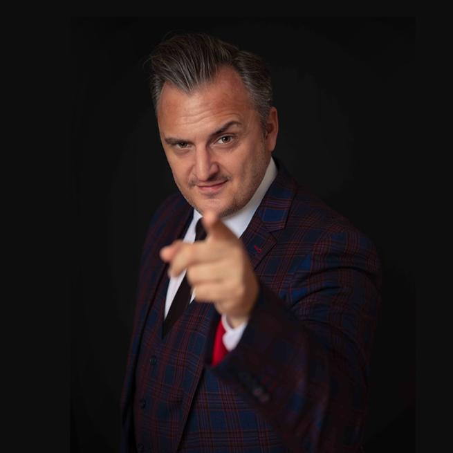 Matt Daniel-Baker in a suit with a blue and red tartan-like suit including a waistcoat and tie, pointing at the camera.