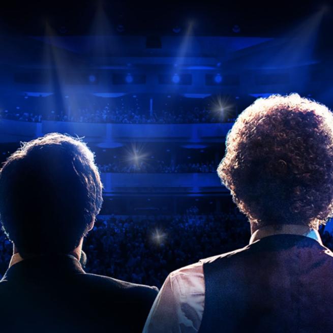 Simon and Garfunkel have their backs to the camera as they face a full theatre. The lights shine on them with a blue colour filling the room.