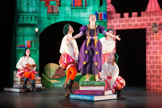 A girl in a princess pressed stood on top of three large books with her arms stretched wide. The background is castle-like. 