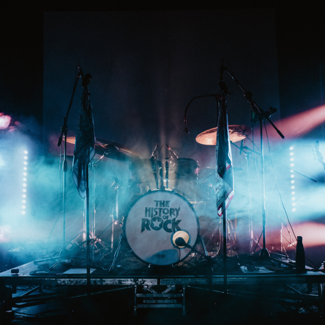 Drum kit on stage with stage lights on it. On the bass drum it says 'The History of Rock'.