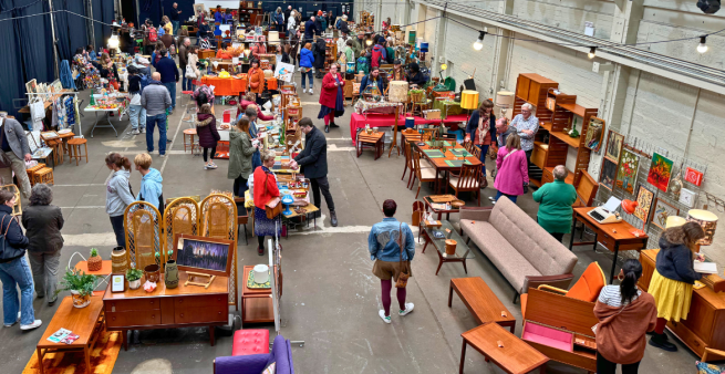 The image overlooks a vintage flea market. There are different items on sale, from tables and chairs to cabinets and decorative homewares. The image shows visitors walking around the market and browsing the different stalls.