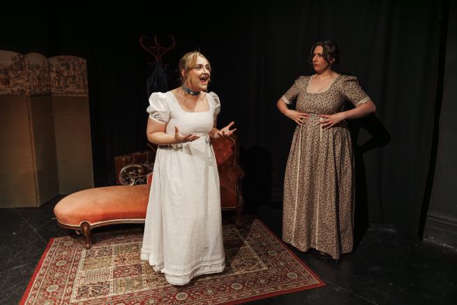 Two women on stage wearing period clothing. One looks excited, the other looks perplexed.