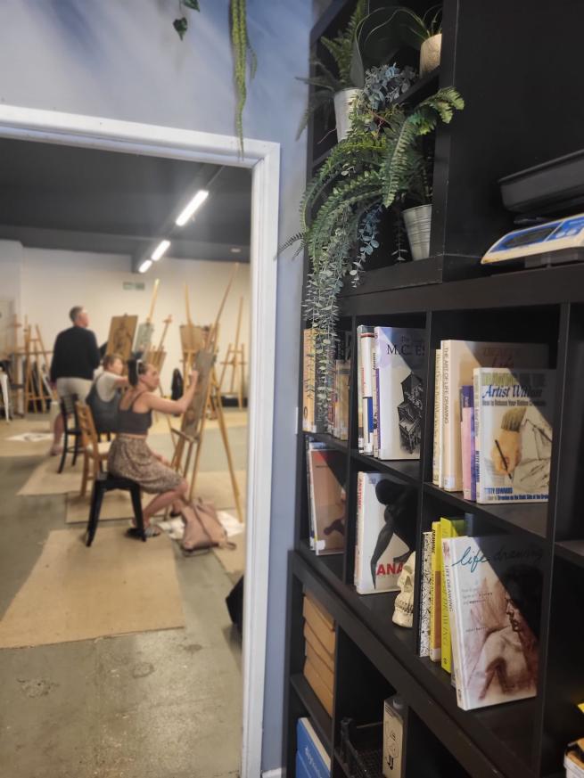 Entrance to an art studio, with bookshelves displaying art books on the right, and people sat and standing at their easels, drawing. 