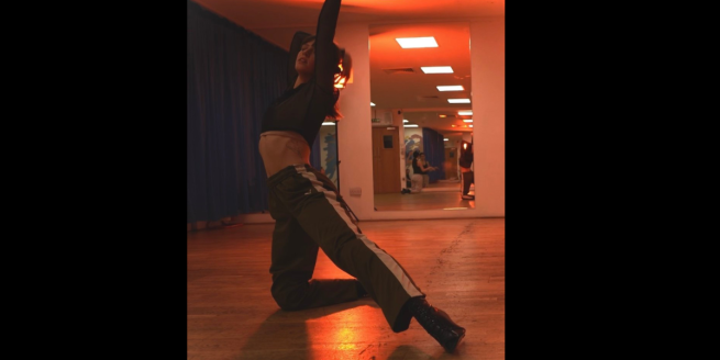 Solo female dancer in a studio, with orange lighting. She is wearing green tracksuit trousers with a white strip down the outside centre of the leg,a green long sleeved cropped top and black lace heels. She is positioned on her knee with her left leg out straight ad arms up, one placed on here head. 