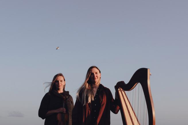 Two woman smiling at the camera. One is holding a harp.