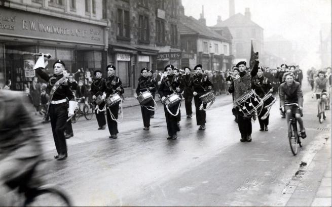 Black and white image of a marching band playing their instruments walking down the street.