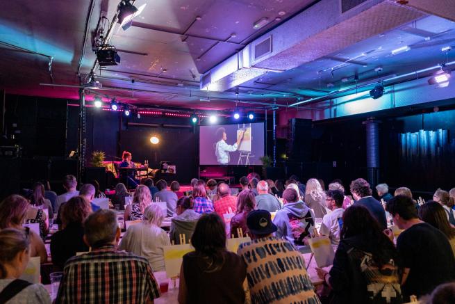 People follow a Painting lesson hosted by Rabbey following a Bob Ross Episode on a big screen