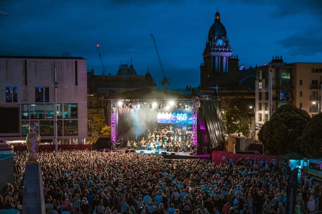 Image of the 80s Classical stage in Millennium Square. In the foreground the concert attendees can be seen with the stage lit at the back. The Leeds Town Hall building can be seen in the background. 