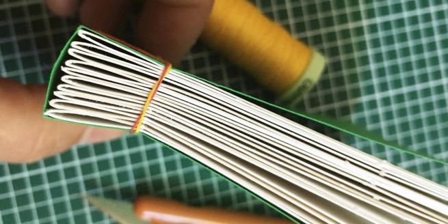 Close up shot of spine of book held between fingers. Thread and cutting mat are in the background.