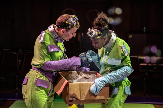 Two people wearing green and purple jumpsuits, looking intently into a cardboard box