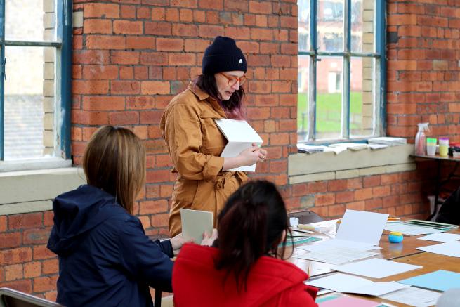 workshop facilitator stands in old industrial space in front of table with paper laid out. 2 participants watch on. 