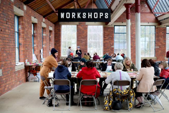 kim stands at the erhad of a table in an old indusrial space. people are sat around the table a 'workshop' sign hangs above their head. 