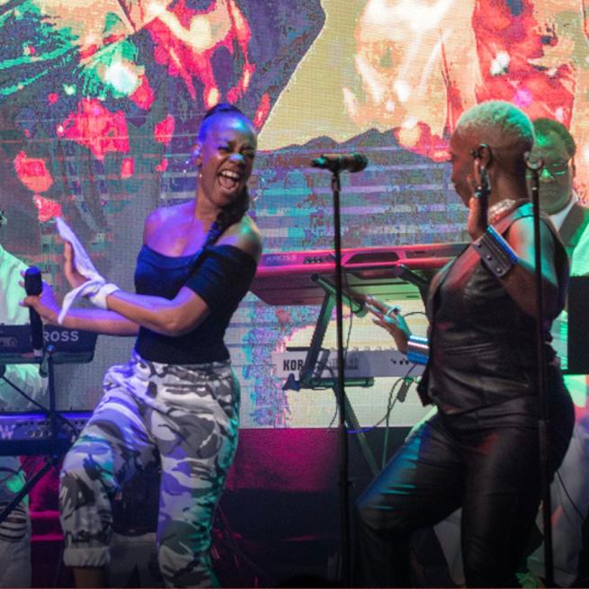 Two singers mid-show in front of their band and a projection screen.