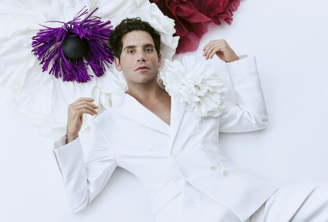 Image of Mika dressed in a white suit. The photograph is taken on a white background with colourful details at the top. 