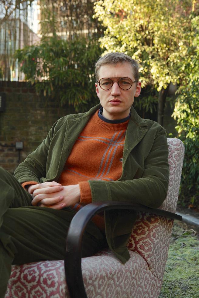 Image of author sitting in a garden with right leg crossed over left.  Author is looking at camera wearing dark trousers, orange jumper and green jacket.