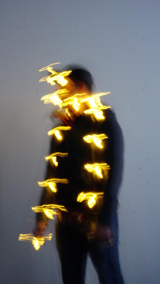 A blurred photograph of a person standing against a plain wall, wrapped in glowing string lights. The movement of the lights creates a dynamic, abstract effect, with streaks of warm yellow light appearing to float around the figure. The person's face and body are obscured due to motion blur, adding a sense of mystery and energy to the image.