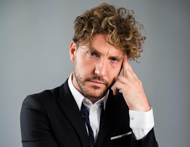 Comedian Seann Walsh dressed in a suit looking thoughtful and holding his hand to his head 