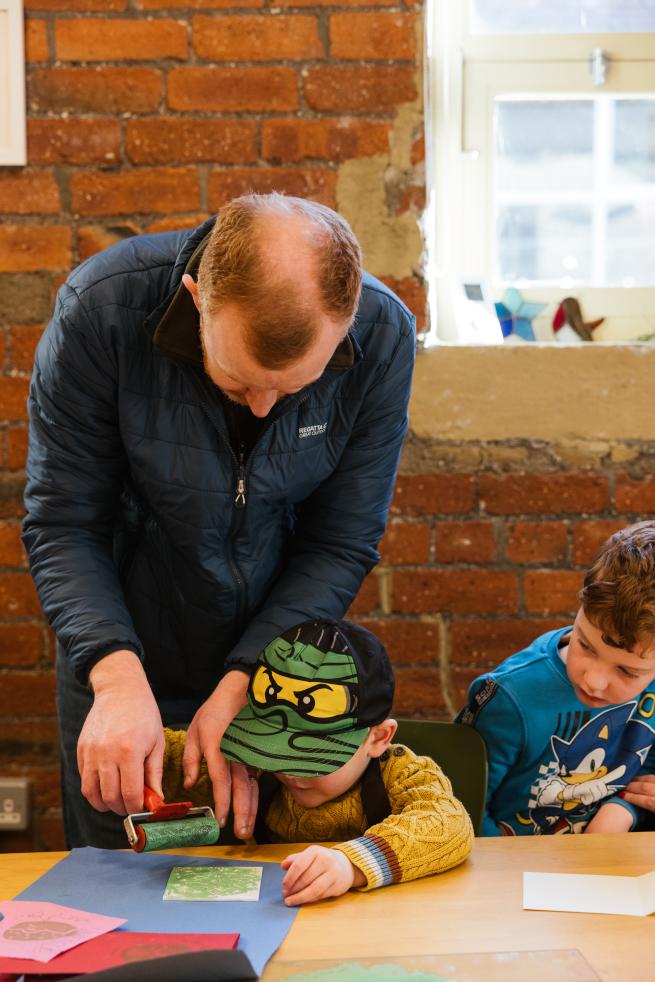 father and child work together on a lino print using a small roller coated in green ink.