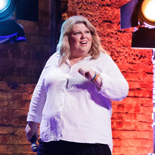 Urzila Carlson smiling on stage holding a microphone down by her right side.