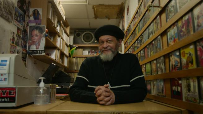 black record store owner owner wearing all black sat in his store surrounded by records