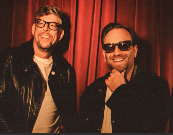 Image of the Black Keys band members smiling in front of a red curtain. 