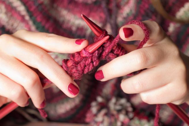 Image shows a lady with red nail polish, holding 2 red knitting needles and wool. 