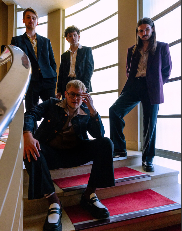 Image of the band members on a staircase. Alex, the band singer is seated on one of the steps whilst the others are around him in the background. They are all looking at the camera. 