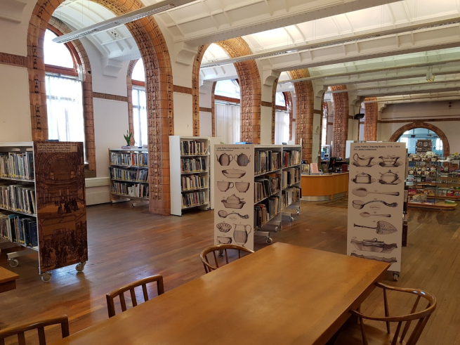 Photo of the Leeds Art Library, showing tiles archways, bookshelves and a long study tables and chairs.